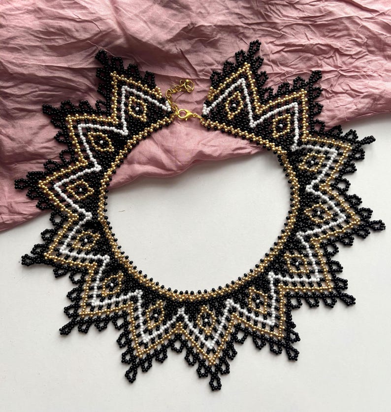 Puede incluir: Un collar de cuentas con un dise&ntilde;o geom&eacute;trico con cuentas negras, doradas y blancas. El collar tiene forma de estrella con un cierre dorado. El intrincado trabajo de cuentas crea un accesorio llamativo.