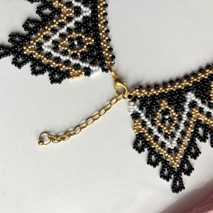 Puede incluir: Un collar de cuentas con un patr&oacute;n geom&eacute;trico en negro, blanco y dorado. El collar tiene un cierre y una cadena dorados para ajustar la longitud. El intrincado trabajo de cuentas crea un dise&ntilde;o texturizado y llamativo.