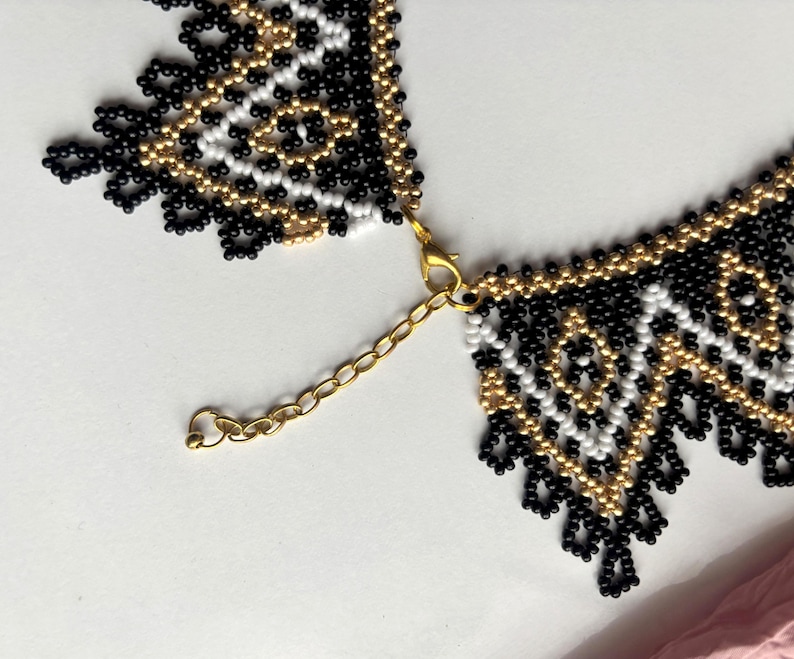 Puede incluir: Un collar de cuentas con un patr&oacute;n geom&eacute;trico en negro, blanco y dorado. El collar tiene un cierre y una cadena dorados para ajustar la longitud. El intrincado trabajo de cuentas crea un dise&ntilde;o texturizado y llamativo.