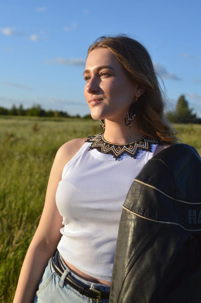 Puede incluir: Una mujer con una camiseta sin mangas blanca, vaqueros azules y una chaqueta de cuero negra. Lleva un collar y pendientes de cuentas con detalles en negro, dorado y blanco. El fondo es un campo y cielo azul.