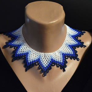 May include: A white and blue beaded necklace with a scalloped edge. The necklace is made of small beads and has a delicate, feminine look.