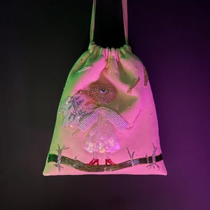 Puede incluir: Una bolsa de cord&oacute;n rosa con un dise&ntilde;o de cuentas de una figura con un ojo grande, que lleva un vestido blanco y zapatos rojos, de pie sobre una rama.
