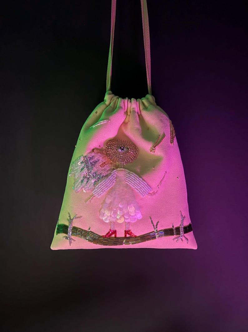 Puede incluir: Una bolsa de cord&oacute;n rosa con un dise&ntilde;o de cuentas de una figura con un ojo grande, que lleva un vestido blanco y zapatos rojos, de pie sobre una rama.