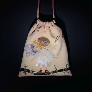 Puede incluir: Una bolsa de cord&oacute;n beige con un dise&ntilde;o de cuentas de una ni&ntilde;a con alas y un ojo grande en su sombrero. La ni&ntilde;a lleva un vestido blanco y zapatos rojos.