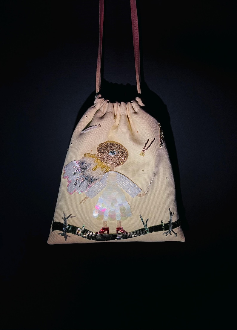 Puede incluir: Una bolsa de cord&oacute;n beige con un dise&ntilde;o de cuentas de una ni&ntilde;a con alas y un ojo grande en su sombrero. La ni&ntilde;a lleva un vestido blanco y zapatos rojos.