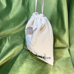 Puede incluir: Una bolsa de cord&oacute;n blanco con un dise&ntilde;o de p&aacute;jaro de cuentas. El p&aacute;jaro tiene un cuerpo azul, alas marrones y un ojo grande. La bolsa tiene una correa blanca larga.