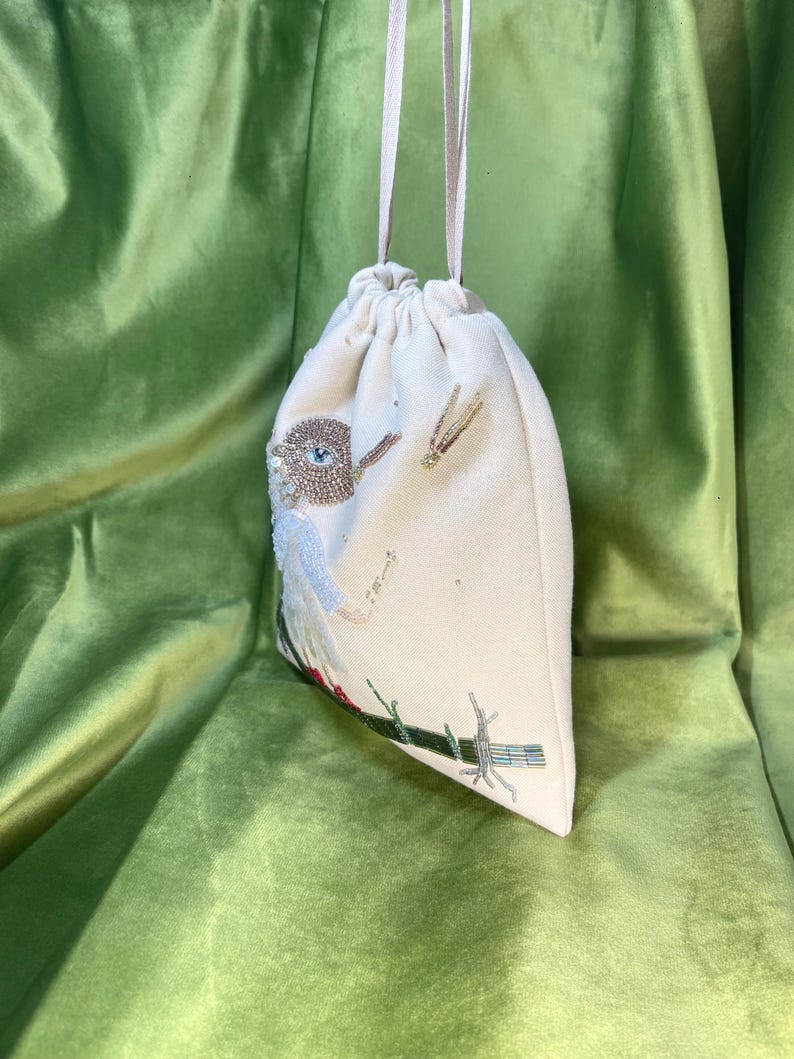 Puede incluir: Una bolsa de cord&oacute;n blanco con un dise&ntilde;o de p&aacute;jaro de cuentas. El p&aacute;jaro tiene un cuerpo azul, alas marrones y un ojo grande. La bolsa tiene una correa blanca larga.