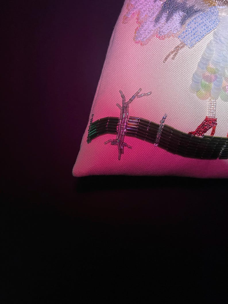 Puede incluir: Primer plano de una almohada bordada en rosa y blanco con un dise&ntilde;o de cuentas de un &aacute;rbol y una figura con zapatos rojos.