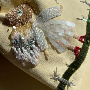 Puede incluir: Una bolsa de tela beige con un dise&ntilde;o de cuentas de un p&aacute;jaro con alas iridiscentes y una rama verde. El p&aacute;jaro lleva un vestido blanco con lentejuelas iridiscentes.