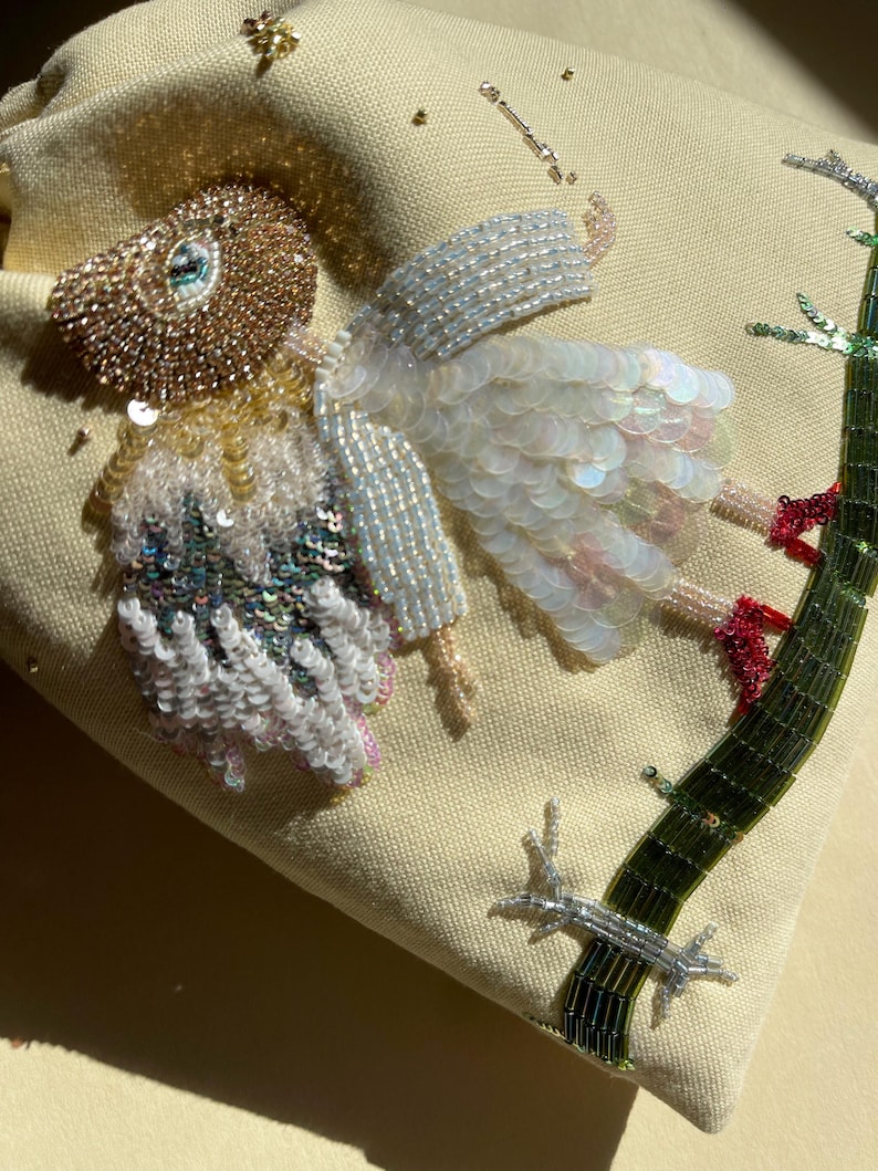 Puede incluir: Una bolsa de tela beige con un dise&ntilde;o de cuentas de un p&aacute;jaro con alas iridiscentes y una rama verde. El p&aacute;jaro lleva un vestido blanco con lentejuelas iridiscentes.