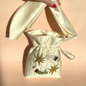 Puede incluir: Una bolsa de cordón de satén blanco con un diseño acolchado y un asa larga de satén. La bolsa está decorada con estrellas y estrellas fugaces bordadas.