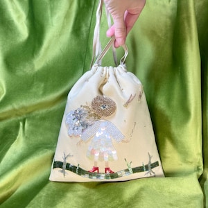 Puede incluir: Una bolsa de cord&oacute;n de color crema con un dise&ntilde;o de cuentas de una ni&ntilde;a con un vestido blanco y zapatos rojos. La ni&ntilde;a est&aacute; de pie sobre una rama verde con detalles de purpurina dorada.