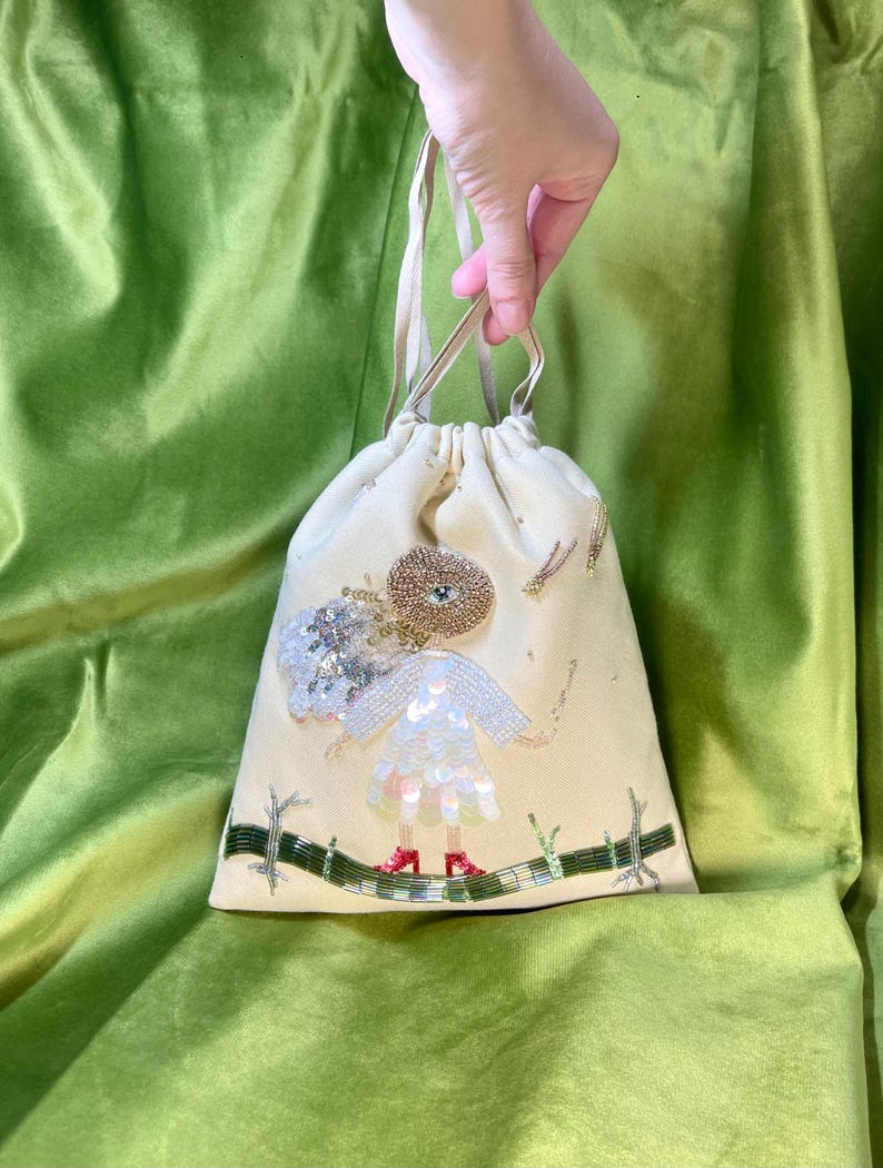 Puede incluir: Una bolsa de cord&oacute;n de color crema con un dise&ntilde;o de cuentas de una ni&ntilde;a con un vestido blanco y zapatos rojos. La ni&ntilde;a est&aacute; de pie sobre una rama verde con detalles de purpurina dorada.