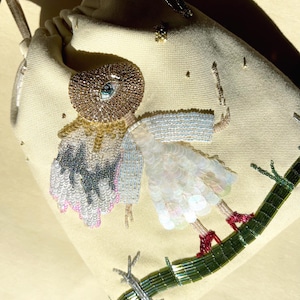 Puede incluir: Una bolsa de cord&oacute;n beige con un dise&ntilde;o de cuentas de una figura con un ojo grande, que lleva un vestido blanco y zapatos rojos, de pie sobre una rama de cuentas verdes.