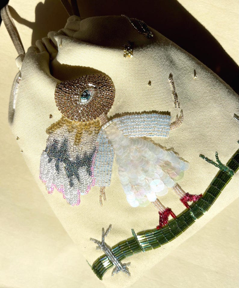 Puede incluir: Una bolsa de cord&oacute;n beige con un dise&ntilde;o de cuentas de una figura con un ojo grande, que lleva un vestido blanco y zapatos rojos, de pie sobre una rama de cuentas verdes.