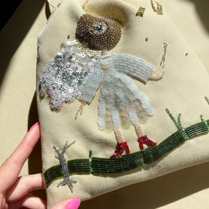 Puede incluir: Una bolsa de cord&oacute;n beige con un dise&ntilde;o de cuentas de una persona con un ojo grande, vistiendo un vestido blanco y zapatos rojos. La persona est&aacute; de pie en un camino de cuentas verdes.