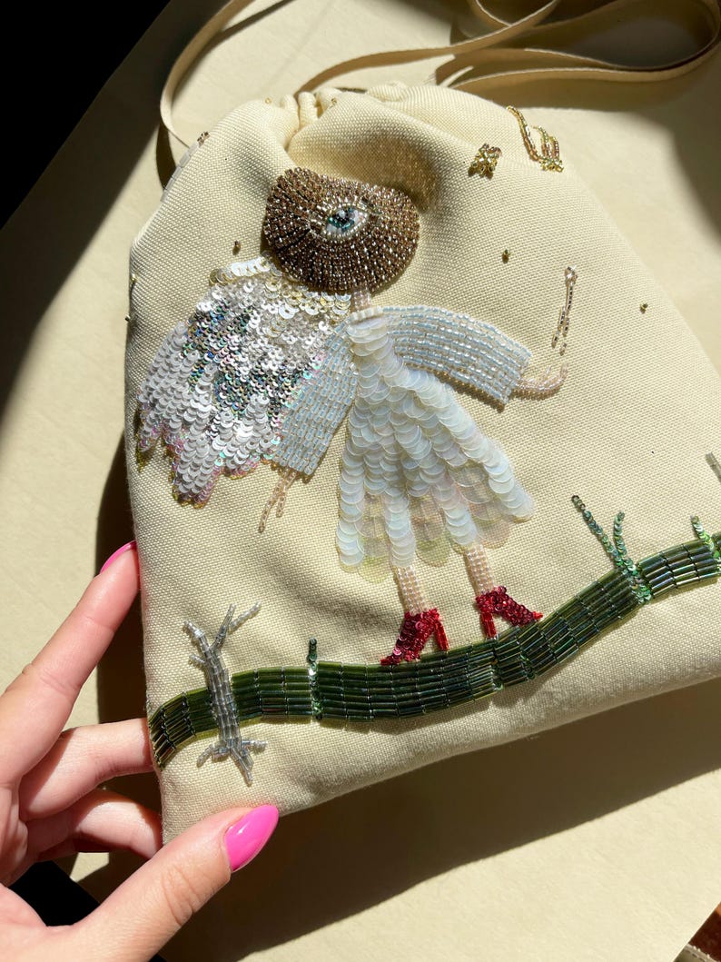 Puede incluir: Una bolsa de cord&oacute;n beige con un dise&ntilde;o de cuentas de una persona con un ojo grande, vistiendo un vestido blanco y zapatos rojos. La persona est&aacute; de pie en un camino de cuentas verdes.