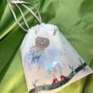 Puede incluir: Una bolsa de cord&oacute;n blanco con un dise&ntilde;o de cuentas de una ni&ntilde;a con un vestido de lentejuelas y zapatos rojos. La bolsa tiene un cord&oacute;n blanco y est&aacute; decorada con cuentas y lentejuelas.