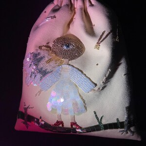 Puede incluir: Una bolsa de cord&oacute;n rosa claro con un dise&ntilde;o de cuentas de una persona con un ojo grande, que lleva un vestido azul y zapatos rojos. La persona est&aacute; de pie sobre una rama de cuentas.