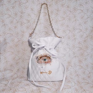 Puede incluir: Bolso blanco acolchado con cordón de ajuste y correa de cadena dorada. El bolso tiene un diseño de ojo con cuentas y un dije de llave dorada.