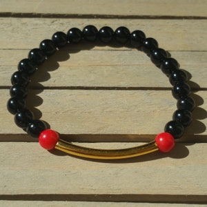 Op de afbeelding: Een zwarte kralen armband met twee rode kralen en een gouden staaf accent.