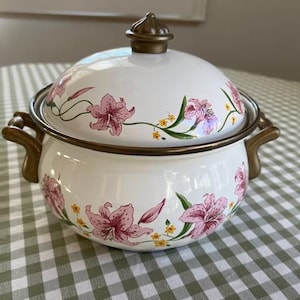 May include: White enamel pot with lid, adorned with pink lily floral design. Features gold-toned handles and rim. A decorative kitchenware item, perfect for serving or display.