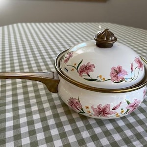 May include: White enamel saucepan with a lid, featuring a floral design in pink and green. The pan has a gold-toned handle and rim. A decorative kitchenware item.