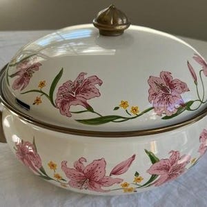 May include: Cream-colored enamelware casserole dish with lid, adorned with pink lily floral pattern and gold-tone handles. The lid is topped with a gold finial. A decorative kitchenware item.