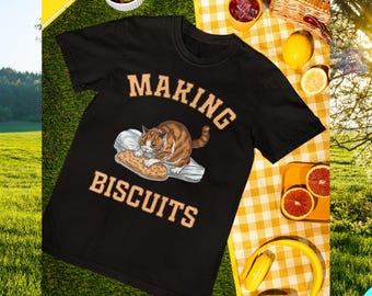 May include: Black t-shirt with the words "MAKING BISCUITS" in tan lettering. A cartoon cat is kneading a biscuit on a pillow. The image is set on a yellow checkered picnic blanket with strawberries, fruit, and headphones.