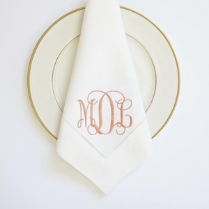 May include: A white linen napkin with a pink embroidered monogram "MDE" on a white plate with a gold rim.