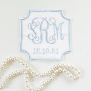 May include: A white fabric patch with a blue border and a monogram of the letters "SRM" in a cursive font. The date "12.10.21" is embroidered below the monogram. A strand of white pearls is lying on a white surface next to the patch.
