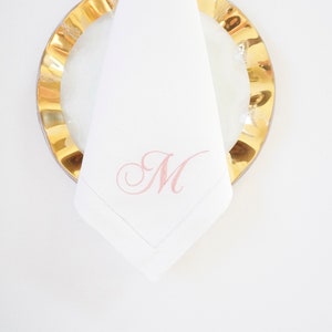 May include: White linen napkin with a stitched border and a delicate pink embroidered letter "M". The napkin is placed on a gold-rimmed plate, creating an elegant and sophisticated table setting.