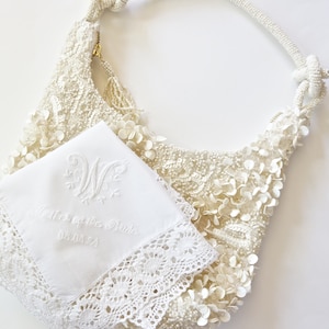 May include: Ivory beaded hobo bag with floral embellishments and a knotted handle. A white handkerchief with lace trim and embroidered text reading "Mother of the Bride" and the date 08.14.24 is next to the bag.