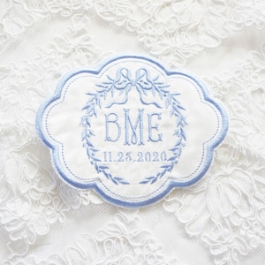 May include: A white embroidered patch with a blue border. The patch features a wreath of leaves, a bow, and the initials "BME" with the date "11.25.2020" below.