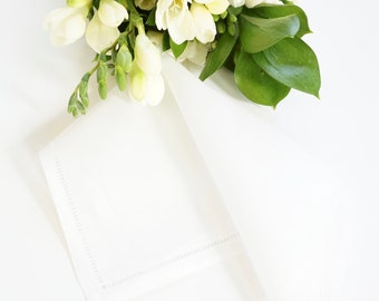 May include: A white linen napkin with a delicate embroidered border. The napkin is folded and placed on a white surface, with a bouquet of white flowers and green leaves in the background.