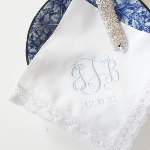 May include: White handkerchief with a blue embroidered monogram and the date 12.31.21. The handkerchief has a white lace trim.