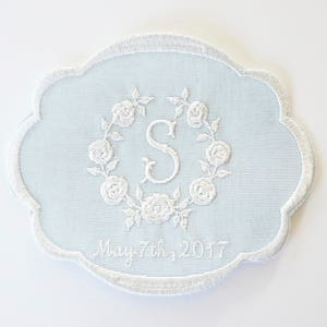 May include: A light blue embroidered cloth coaster featuring a white floral wreath with the letter "S" in the centre. The date "May 7th, 2017" is embroidered below the wreath.