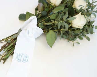 May include: A white bouquet of white roses and greenery tied with a white satin ribbon with the monogram "M.O.E." embroidered in blue thread.