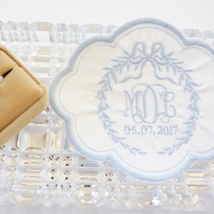 May include: A white and blue embroidered ring bearer pillow with the initials "M.O.E." and the date "05.07.2017". The pillow is on a clear glass tray with a ring box next to it.