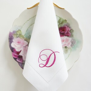 May include: White linen napkin with a pink embroidered letter "D" on a floral patterned plate.