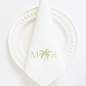 May include: White linen napkin with a green palm tree and the letters "M" and "R" embroidered on it. The napkin is folded and placed on a white plate with a beaded edge.