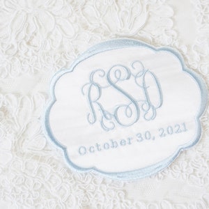 May include: A white fabric patch with a scalloped edge and light blue embroidery. The center features a decorative monogram with the letters RSD, and the text October 30, 2021 is embroidered in light blue below the monogram on the white surface.