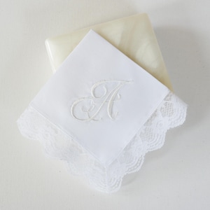 May include: A square white handkerchief featuring a delicate white lace border with a scalloped edge. The center of the fabric is embroidered with a single, elegant script letter A in white thread, creating a monochromatic and detailed textile design.