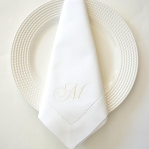May include: A white linen napkin with an embroidered "M" rests on a white plate with a textured rim. The napkin is folded into a triangle shape, showcasing the elegant monogram detail. Ideal for formal dining.