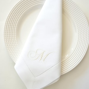 May include: A white linen napkin with an embroidered letter "M" rests on a white plate. The napkin is folded, showcasing the embroidered initial and a delicate stitched border. The plate has a textured rim.