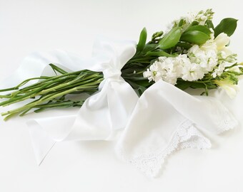 May include: A bouquet of white flowers, including stock and freesia, is tied with a white satin ribbon. A white handkerchief with a lace edge is placed near the flowers. The arrangement is set against a white background.
