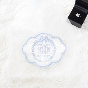 May include: A white fabric pouch featuring a scalloped-edge patch embroidered with light blue thread. The embroidery displays a bow, a laurel wreath, the monogram HGW, and the date 06.19.21. A black ring box containing a square-cut diamond ring sits nearby.