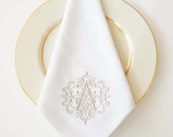 May include: White linen napkin with an embroidered monogram letter "A" in a decorative, ornate design. The napkin is folded and placed on a cream-colored plate with a gold rim, creating an elegant table setting.
