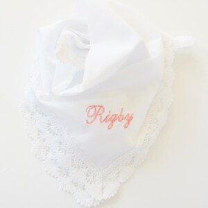 Op de afbeelding: Witte zakdoek met een delicate kanten rand en de naam "Rigby" geborduurd in roze script. De zakdoek is gevouwen, met een kwastje zichtbaar. Een klassiek en elegant accessoire.