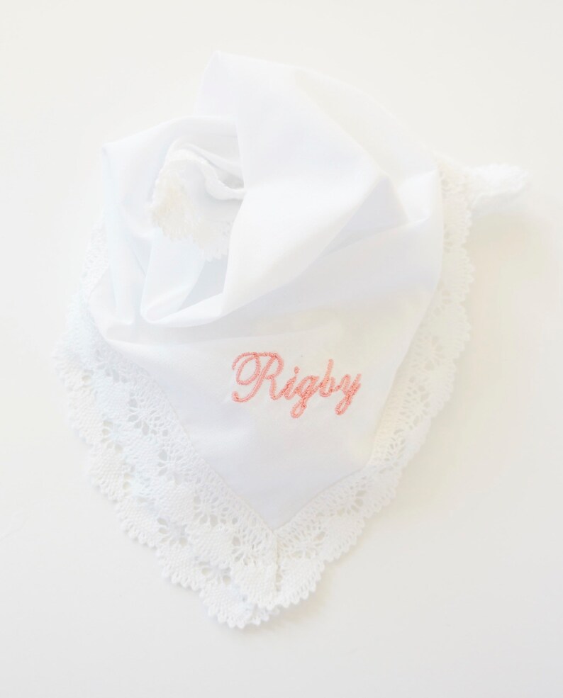 Op de afbeelding: Witte zakdoek met een delicate kanten rand en de naam "Rigby" geborduurd in roze script. De zakdoek is gevouwen, met een kwastje zichtbaar. Een klassiek en elegant accessoire.